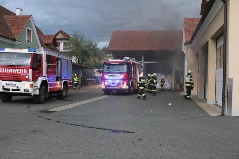 Feuerwehrleute kümmern sich um einen Brand in einem Gebäude. Der Rauch kommt aus dem Gebäude. Zwei Feuerwehrwagen sind am Straßenrand geparkt.