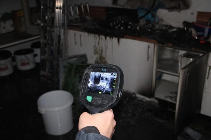 Eine Person hält eine Wärmebildkamera und zeigt eine Ansicht von innen aus einem beschädigten Gebäude. Der Raum ist mit Schutt gefüllt, und weiße Eimer sind auf dem Boden verstreut.
