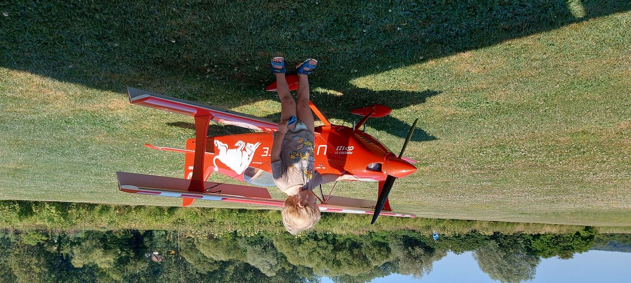 A young boy is hanging upside down from the underside of a red airplane on a grassy field.