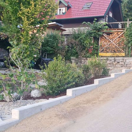 Ein Haus mit rotem Dach steht hinter einer Steinmauer, mit einer Einfahrt und einem Garten mit Sträuchern und einem kleinen Baum.