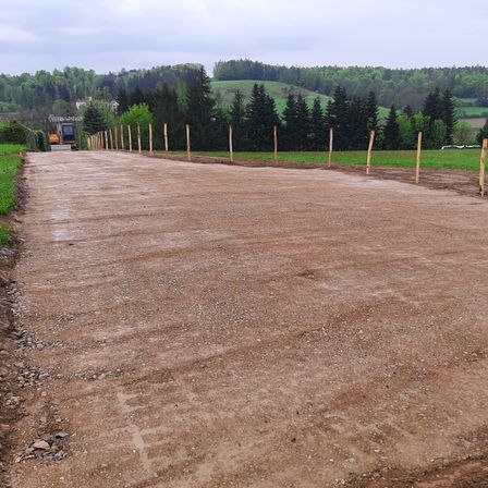 Eine unbefestigte Straße mit einem Baufahrzeug in der Ferne, umgeben von Holzpfählen und grünen Feldern.