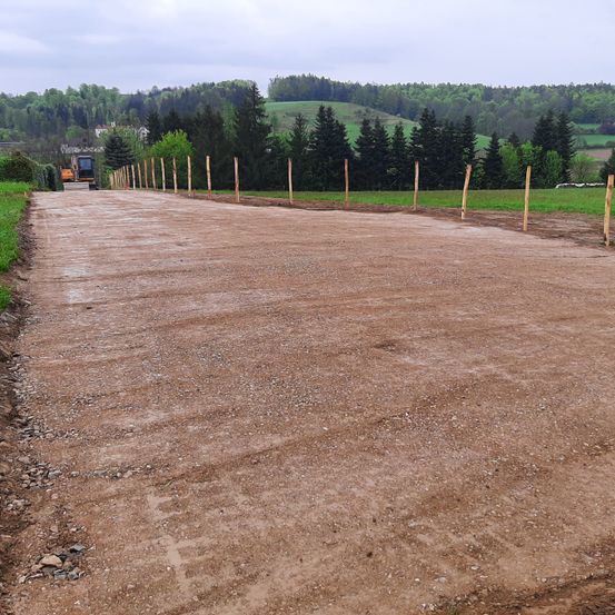 Eine unbefestigte Straße mit einem Baufahrzeug in der Ferne, umgeben von Holzpfählen und grünen Feldern.