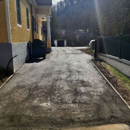 Eine Auffahrt führt zu einem Haus mit gelber Wand und schwarzem Tor. Hinter dem Haus befindet sich eine Garage. Die Straße ist leer. Bäume und Gras befinden sich an den Seiten.
