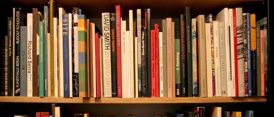 Ein hölzerne Bücherregal gefüllt mit zahlreichen Büchern in verschiedenen Größen und Farben. Titel beinhalten 'SITE', 'David Smith' und 'Ruefino Tamayo'. Die Bücher sind ordentlich in Reihen angeordnet.