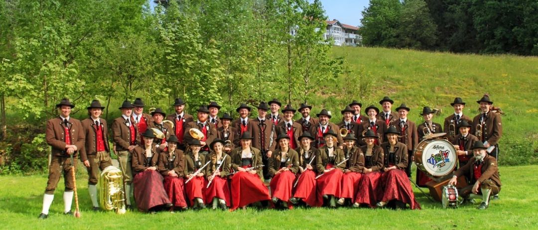 Eine große Gruppe von Musikern in traditioneller Kleidung, einige mit Musikinstrumenten, posiert für ein Foto auf einem Grashügel. Bäume und ein Gebäude im Hintergrund.