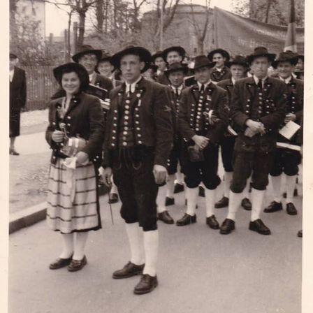 Eine Gruppe von Personen in traditioneller Kleidung marschiert in einem Umzug. Eine Frau in einem Rock hält einen Preis in der Hand.