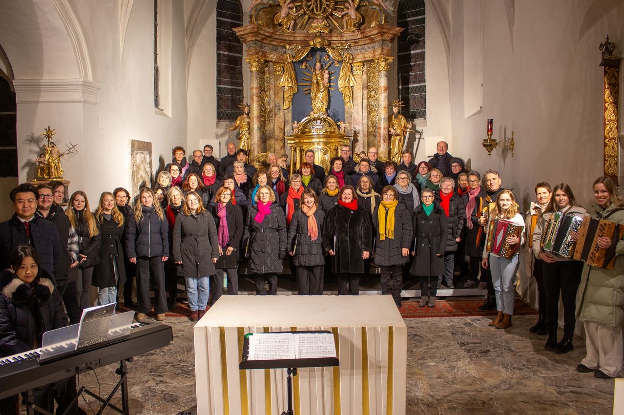 Ein Chor steht vor einem Altar in einer Kirche. Die Mitglieder tragen Winterkleidung und halten Notenblätter. Dahinter befindet sich ein kunstvoller Altar mit Statuen.