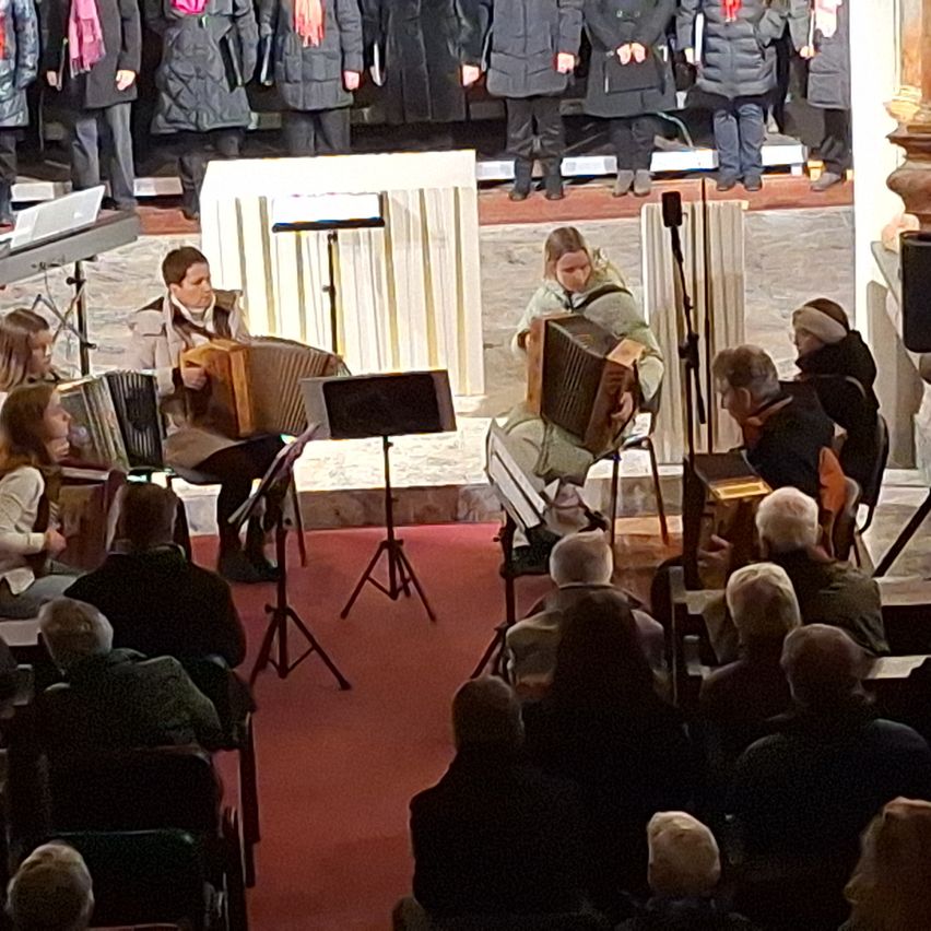 Eine Gruppe von Menschen spielt Akkordeons in einer Kirche. Ein Publikum sitzt auf Stühlen vor ihnen. Hinter den Musikern steht ein Chor auf der Bühne.