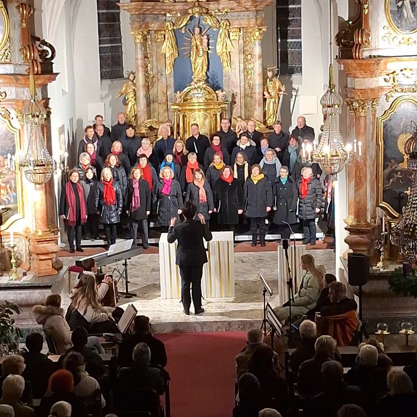 Ein Chor tritt in einer Kirche mit verzierten Säulen, goldenen Statuen und einer Kristallleuchte auf. Der Dirigent führt auf einer Bühne mit rotem Teppich. Ein Publikum sitzt davor.