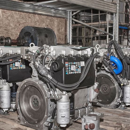 Mehrere industrielle Motoren mit verschiedenen Komponenten und Etiketten sind auf einem Holzpalette in einem Lagerhaus angeordnet. Einige Motoren haben blaue und schwarze Schläuche, während andere metallische Teile aufweisen. Der Hintergrund zeigt eine Ziegelwand und Metallstrukturen.