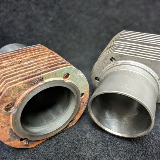 Zwei Motorteile, eines rostig mit einer abgenutzten Oberfläche und das andere poliert und sauber, sind nebeneinander auf einem dunklen Hintergrund ausgestellt.