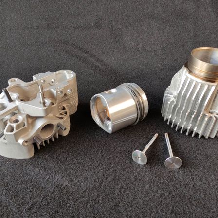 Eine Sammlung von Motorkomponenten, darunter ein Zylinderkopf, ein Kolben und zwei Kolbenringe, liegen auf einer grauen Oberfläche.