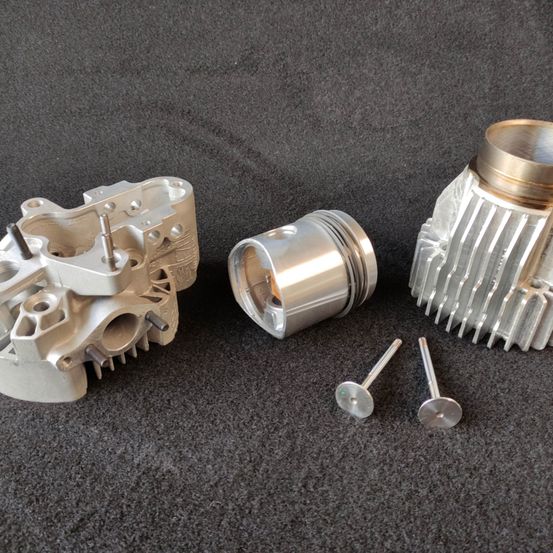 Eine Sammlung von Motorkomponenten, darunter ein Zylinderkopf, ein Kolben und zwei Kolbenringe, liegen auf einer grauen Oberfläche.