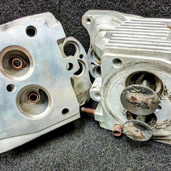 Zwei zerlegte Metall-Motorbauteile mit verschiedenen Löchern und Schrauben auf einer grauen Oberfläche. Eines hat ein kreisförmiges, korrodiertes Stück.