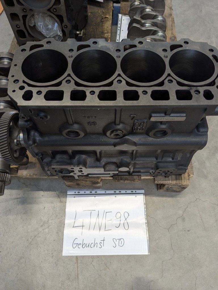 Ein großer Motorblock mit einem Schild, auf dem '4TNE98 Gebuchst STD' steht, ist auf einem Holzpalette ausgestellt. Der Motorblock ist schwarz und grau und verfügt über vier Zylinderköpfe.