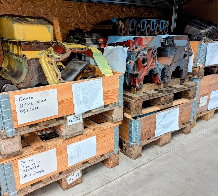 Ein Lager mit mehreren Holzkisten, die auf Paletten gestapelt sind, jede mit spezifischen Codes und Beschreibungen beschriftet. Die Kisten enthalten verschiedene Teile, einschließlich Motorkomponenten. Eine Kiste hat das Etikett 'Deutz BF4L1011A 7920683'.