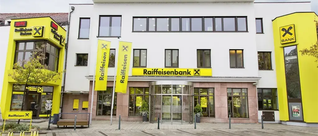 Ein weißes Gebäude mit mehreren Fenstern hat ein Schild mit der Aufschrift Raiffeisenbank über dem Eingang. Zwei gelbe Banner stehen vor dem Gebäude. Draußen gibt es eine Bank und einen Topf mit Pflanzen.