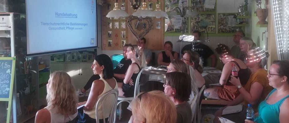 Eine Gruppe von Menschen sitzt in einem Raum auf Stühlen und beobachtet eine Präsentation auf einer Projektionsleinwand. Das Thema ist Tierpflege.