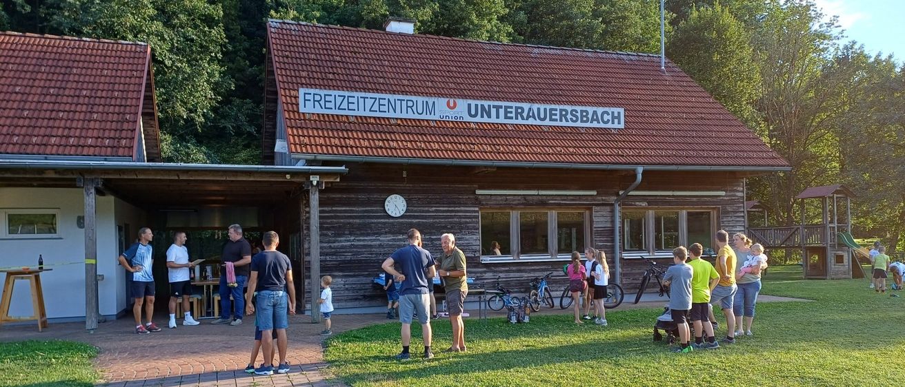 Eine Gruppe von Menschen steht vor einem Gebäude mit einem Schild, das Freizeitzentrum Unterauersbach besagt. Fahrräder sind vor dem Gebäude geparkt. Bäume sind im Hintergrund.