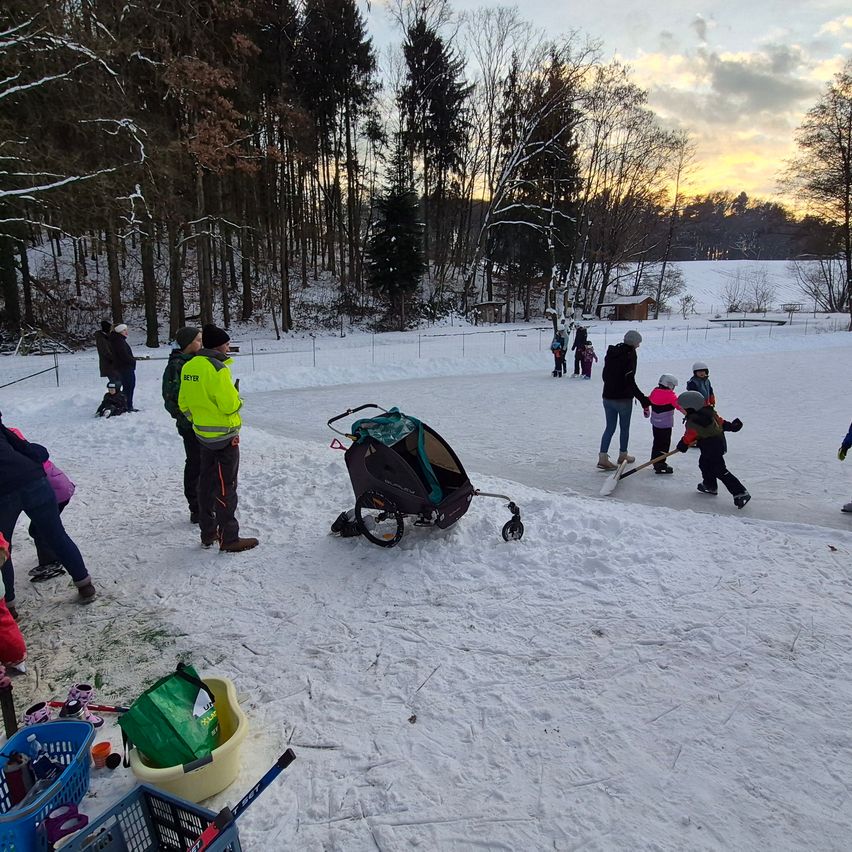 Ein verschneiter Außenbereich mit mehreren Menschen, einige spielen Eislaufen und andere stehen herum. Der Bereich ist von Bäumen umgeben.