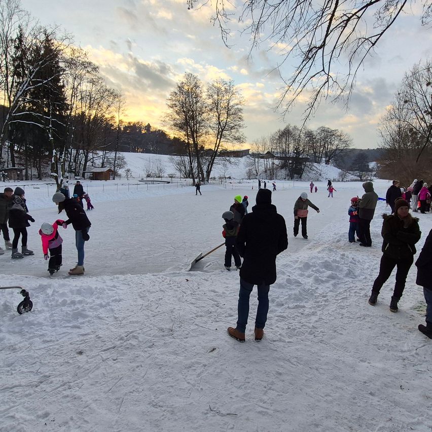 Eine verschneite Landschaft mit Menschen, die auf einer Eisbahn Schlittschuh laufen, stehen und herumlaufen. Bäume und ein Zaun umgeben das Gebiet, mit einem Blick auf ein Haus in der Ferne.