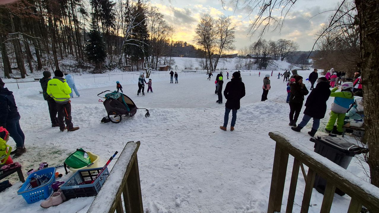 Eine Eislaufbahn in einem verschneiten Gebiet, umgeben von Bäumen. Mehrere Personen sind beim Eislaufen, während andere herumstehen. Ein Kinderwagen ist in der Nähe.