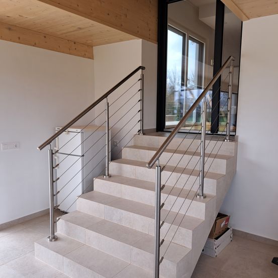 Eine moderne Treppe mit weißen Fliesen und Metallhandläufen. Die Decke ist aus Holz und oben befindet sich eine Glastür. Ein Kühlschrank steht neben der Treppe.