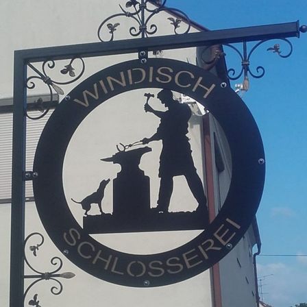 Ein rundes Schild zeigt einen Schmied, der auf einen Amboss schlägt, begleitet von einem Hund. Der Text lautet 'WINDISCH SCHLOSSEREI'.
