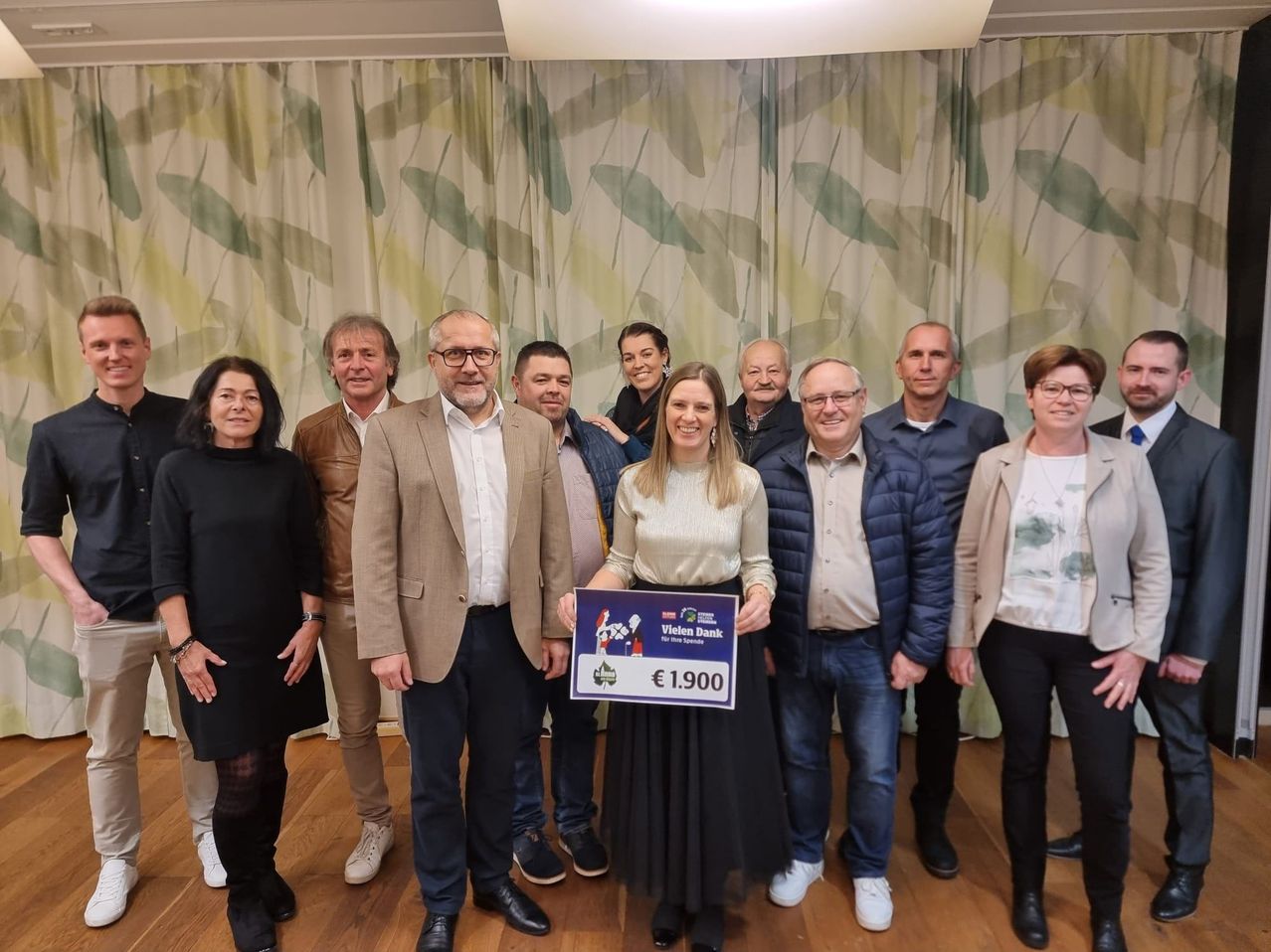 Eine Gruppe von Menschen posiert für ein Foto, während sie einen großen Scheck halten. Die Frau in der Mitte hält den Scheck und lächelt. Der Scheck sagt '€ 1.900'. Hinter ihnen ist ein Vorhang mit einem Blattdesign.
