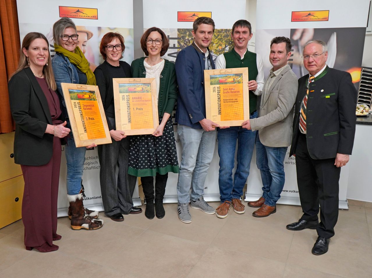 Eine Gruppe von Menschen, sowohl Männer als auch Frauen, steht zusammen und hält gerahmte Urkunden in den Händen. Sie lächeln und posieren für ein Foto. Einige von ihnen tragen Brillen, Stiefel und Schals. Der Mann in der Mitte trägt einen Anzug und eine Krawatte. Hinter ihnen ist ein weißer Banner mit Text und Logos.