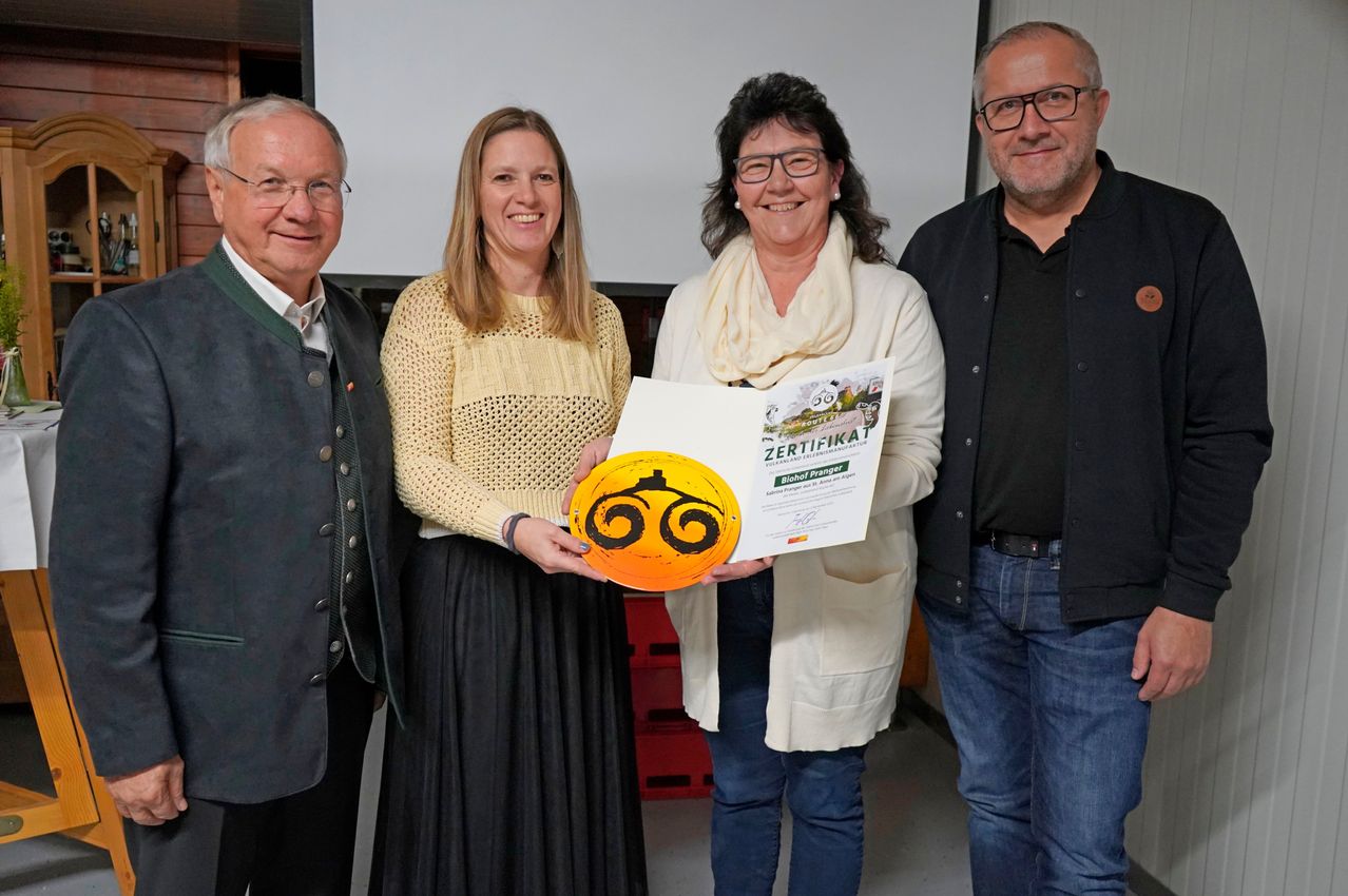 Vier Menschen stehen drinnen zusammen. Eine Frau hält ein Zertifikat und eine runde Plakette. Der Mann rechts trägt eine Brille.