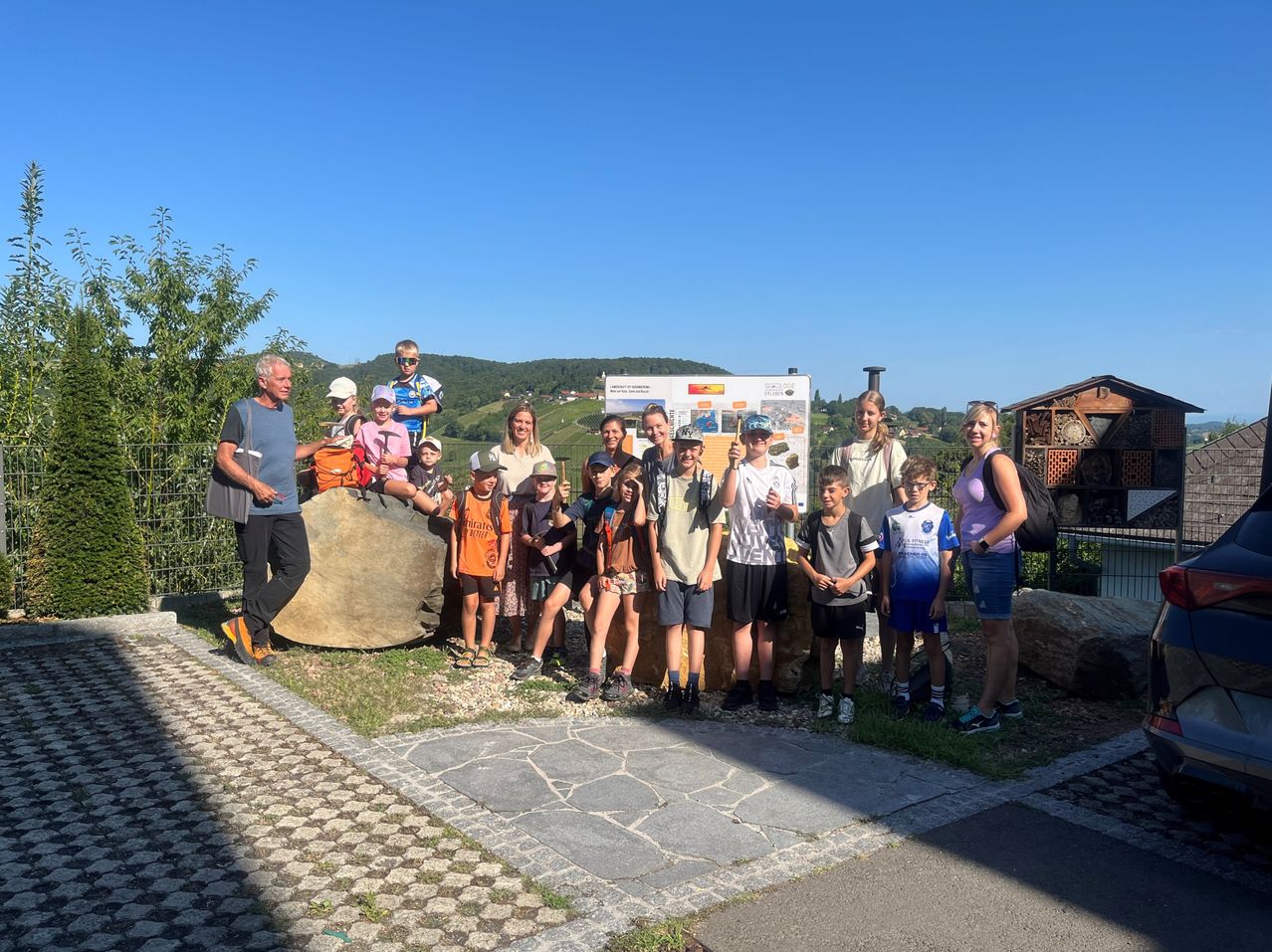 Eine Gruppe von Menschen, einschließlich Kindern, steht vor einem großen Felsen und einem Schild und posiert für ein Foto. Sie sind lässig gekleidet und scheinen auf einer Exkursion zu sein.
