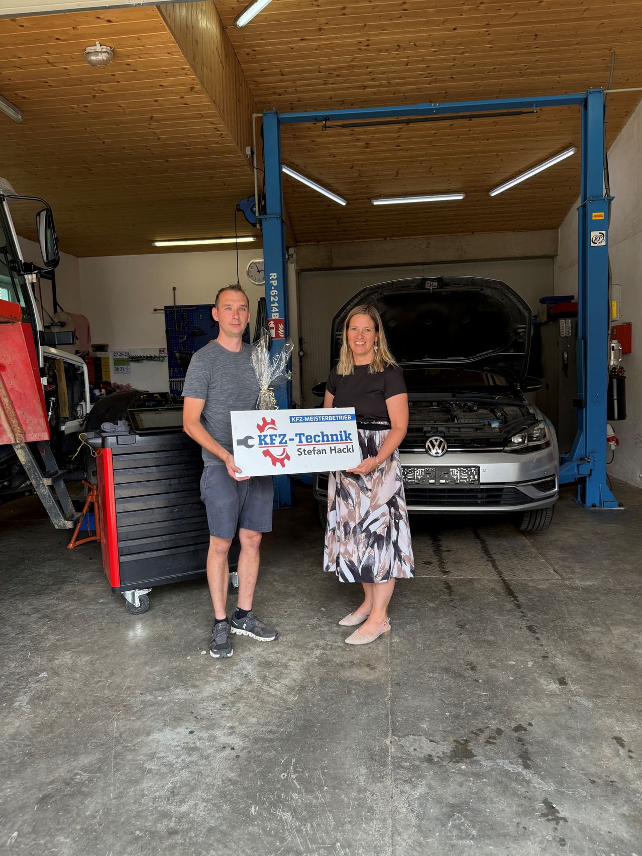 Ein Mann und eine Frau stehen in einer Garage und halten ein Schild mit der Aufschrift KFZ-Technik Stefan Hackl. Dahinter steht ein Auto auf einem Wagenheber.