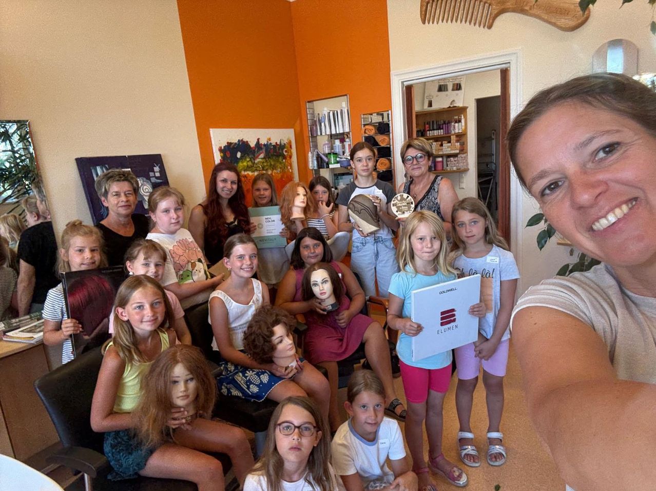 Eine Gruppe junger Mädchen und Frauen posiert für ein Foto in einem Salon mit Mannequinköpfen, einige halten Auszeichnungen und Zertifikate.