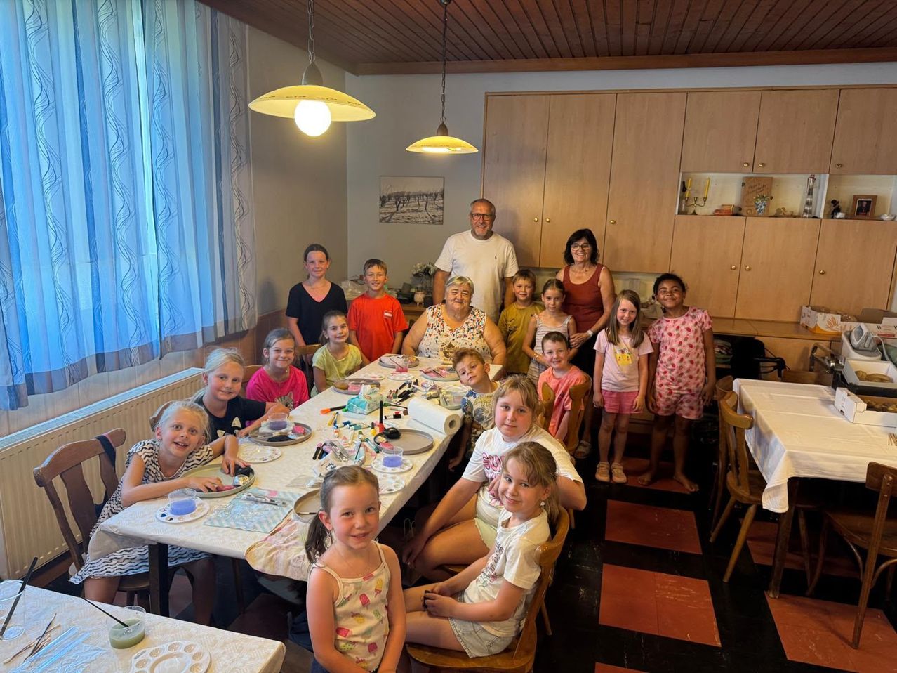 Eine Gruppe von Kindern und Erwachsenen versammelt sich um einen Tisch in einem Esszimmer, lächelt und posiert für ein Foto. Der Tisch ist mit Tellern, Tassen und Malutensilien gedeckt.