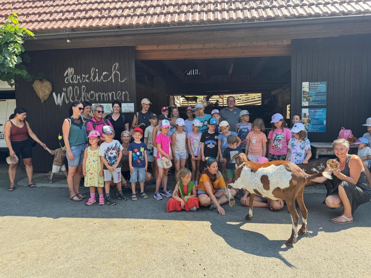 Eine Gruppe von Erwachsenen und Kindern steht vor einem Stall mit Ziegen und lächelt für ein Foto. Der Stall hat ein Schild mit der Aufschrift Herzlich Willkommen.