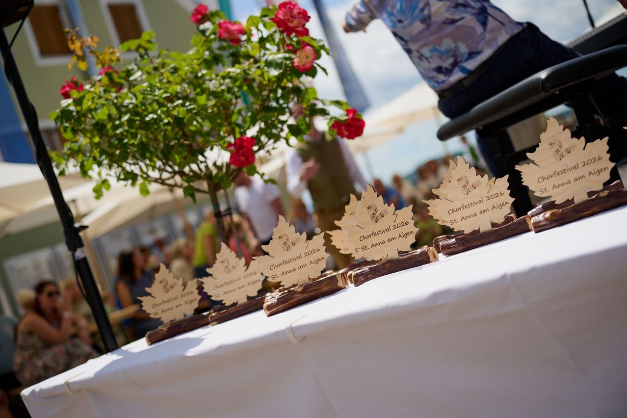 Fünf Holztrophäen mit Ahornblättern sind auf einer weißen Tischdecke angeordnet. Jede Trophäe zeigt den Text 'Chorfestival 2026' und 'St. Anna am Aigen.' Im Hintergrund sind verschwommen Menschen, eine Pflanze mit roten Blumen und Zelte zu sehen.