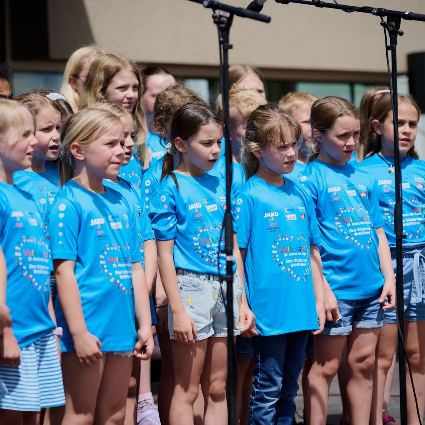 Eine Gruppe junger Mädchen in blauen T-Shirts und Jeansshorts singt in einem Chor, hinter einem Mikrofon vor einem Gebäude stehend.