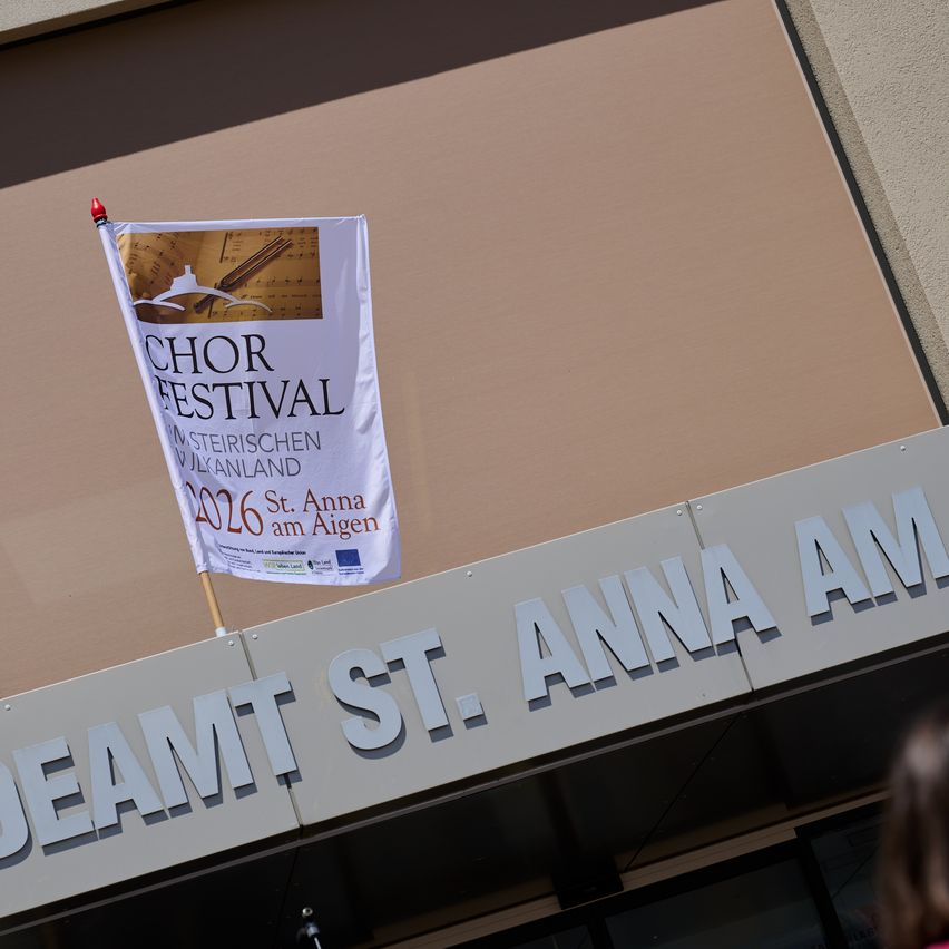 Eine weiße Flagge mit den Worten Chor Festival 2026 ist am Eingang von Deamt St. Anna am Aigen befestigt. Das Gebäude hat eine braune Wand und ein graues Dach.
