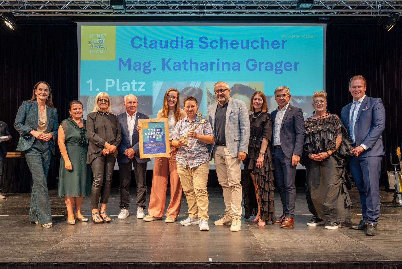 Eine Gruppe von Menschen steht lächelnd auf der Bühne. Eine Person hält einen gerahmten Preis mit dem Text Tierschutz Preis. Hinter ihnen ist eine Leinwand mit Claudia Scheucher Mag. Katharina Grager.