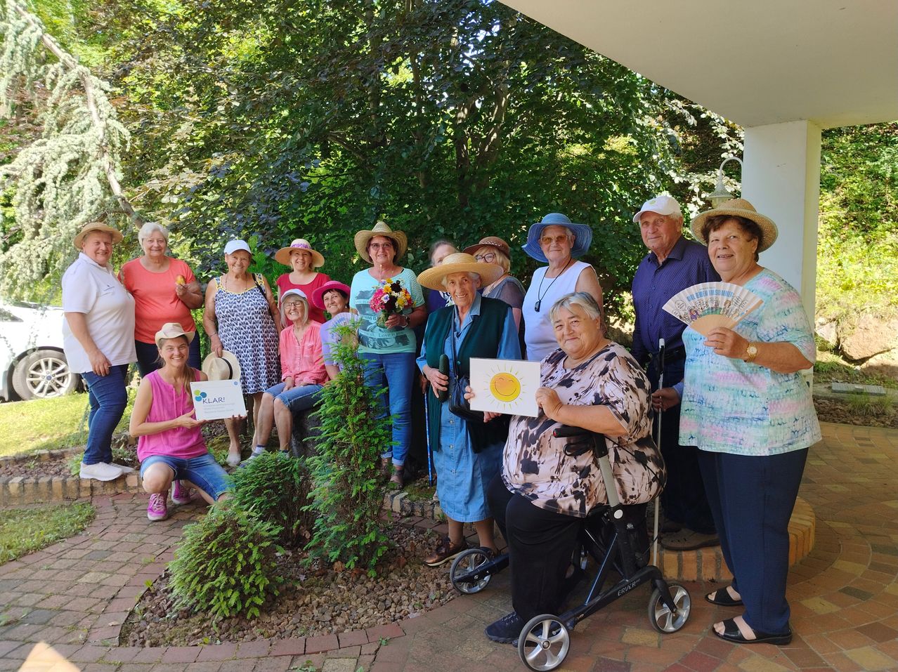 Eine Gruppe älterer Menschen posiert für ein Foto im Garten, einige tragen Hüte und halten Urkunden, eine Frau im Rollstuhl und eine Frau mit einem Fächer.