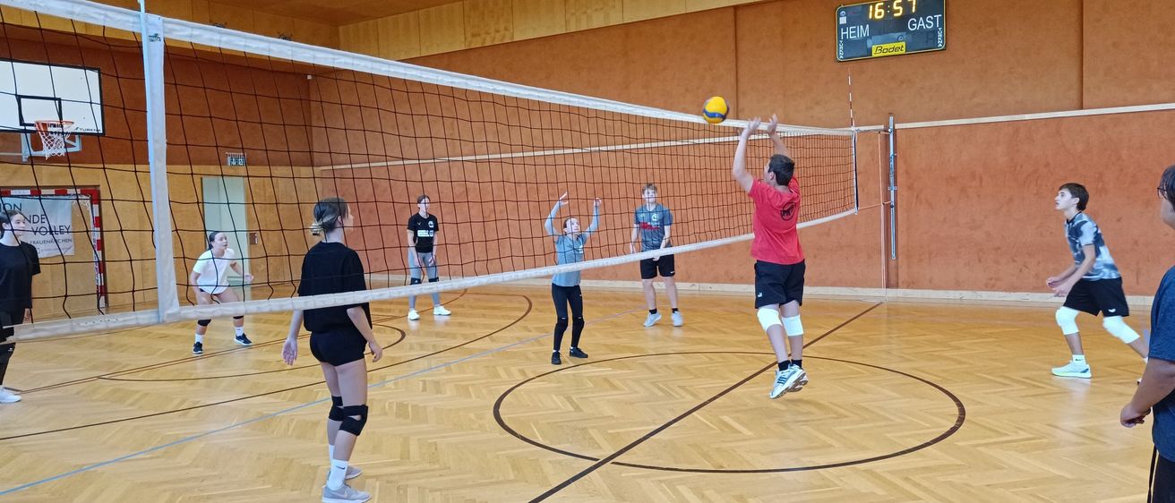 Eine Gruppe von Menschen spielt Volleyball in einer Halle. Ein Spieler springt, um den Ball zu schlagen. Der Platz hat Holzboden und ein Netz.