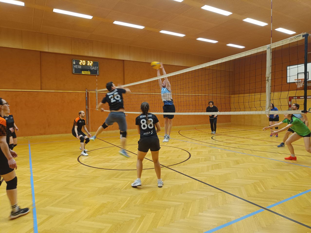 Hallenvolleyballspiel mit Spielern in Trikots. Ein Spieler schlägt den Ball über das Netz, während andere zuschauen. Das Ergebnis steht bei 20-32.