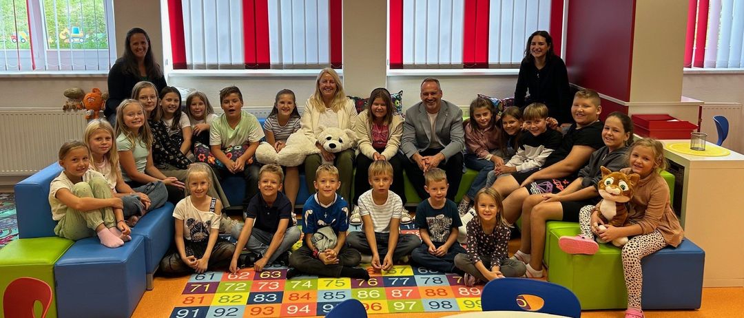 Eine Gruppe von Kindern und Erwachsenen sitzt auf einer Couch im Klassenzimmer, eine Frau hält einen Teddybären und ein Zahlenteppich liegt auf dem Boden.