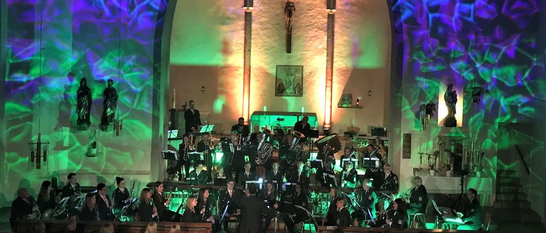 Ein Brass-Band spielt auf einer Bühne, die mit grünen und blauen Lichtern beleuchtet ist. Das Publikum sitzt vor der Bühne. Die Wände sind mit grünen und blauen Lichtern verziert. Ein Kreuz befindet sich an der Wand über der Bühne.