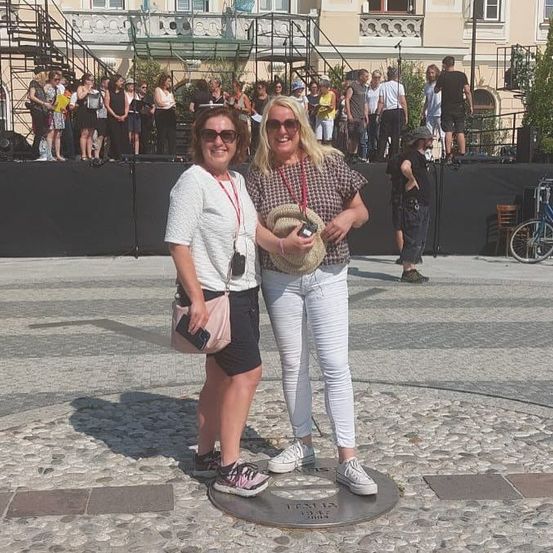 Zwei Frauen stehen vor einer kreisförmigen Plakette mit einem Gebäude und einer Menschenmenge im Hintergrund. Eine trägt eine Sonnenbrille und einen Hut, die andere eine Halskette und eine rosa Tasche.