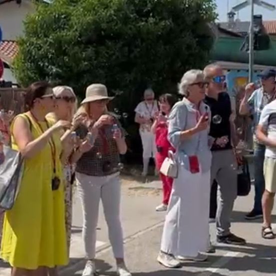 Eine Gruppe von Erwachsenen steht auf einer Straße, einige mit Hüten, Brillen und Taschen. Sie führen Gespräche, möglicherweise Touristen. Bäume und Gebäude sind im Hintergrund.