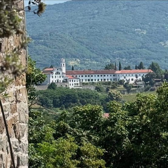 Ein großes weißes Gebäude mit Turm steht auf einem Hügel, umgeben von Bäumen und Bergen.