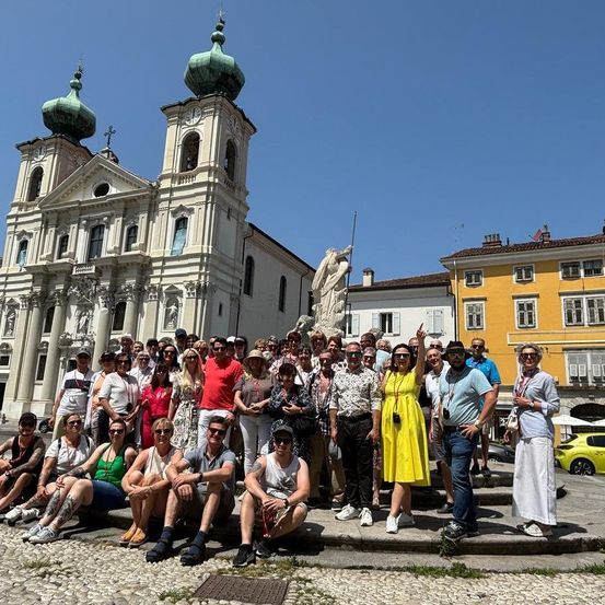 Eine große Gruppe von Touristen posiert an einem sonnigen Tag vor einer Kirche, einige mit Sonnenbrillen und Hüten. In der Mitte steht eine Statue.