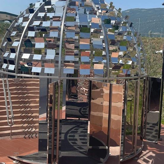 Eine metallische, kuppelförmige Struktur mit zahlreichen reflektierenden Paneelen, die auf einem gefliesten Boden mit einem Eingang steht. Ein Berg und Grünflächen sind im Hintergrund sichtbar.